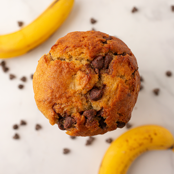View from above of a warm fresh banana chocolate chip muffin atop a plate and garnished with semi sweet chocolate chips and whole bananas.
