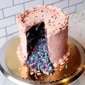 A decadent chocolate cake, iced in pink strawberry buttercream icing with a slice cut out and sprinkles pouring out on to the cake board.