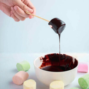 A marshmallow on a stick being lifted away from a bowl of chocolate with the chocolate dripping of the marshmallow.