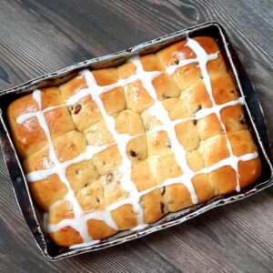 One dozen warm hot cross buns with raisins in a baking pan drizzled with icing in a cross design.