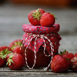 A mason jar filled with fresh strawberry jam covered with a cloth top, that's tied tightly with twine. Strawberries are atop the jar and adorned on the table surrounding the jar.